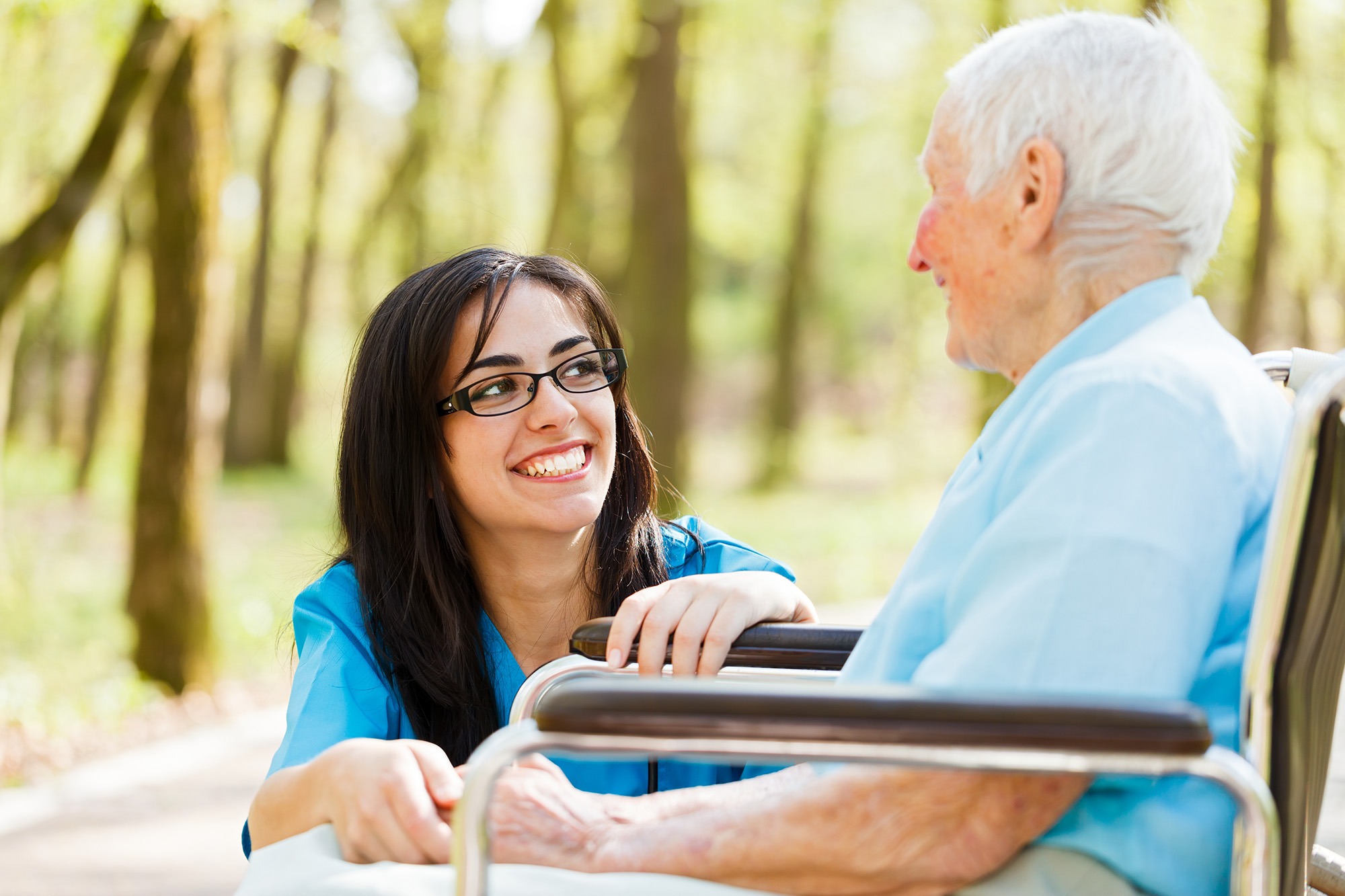 A caregiver smiles warmly at an elderly man in a wheelchair, enjoying a sunny day in a park surrounded by trees, highlighting compassion in caregiving.