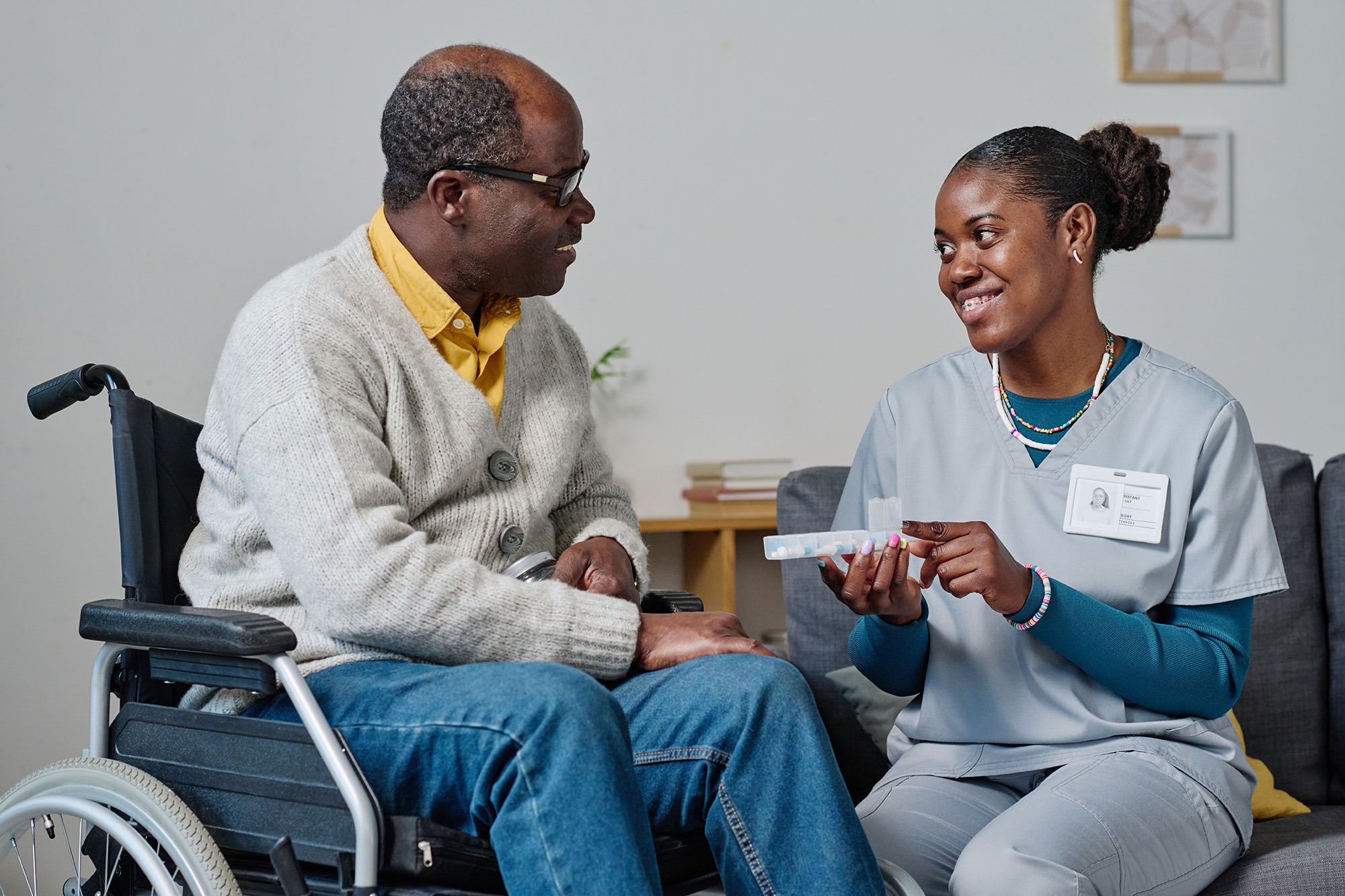 A caregiver in scrubs discusses medication with a smiling man in a wheelchair, highlighting the importance of health support and communication.