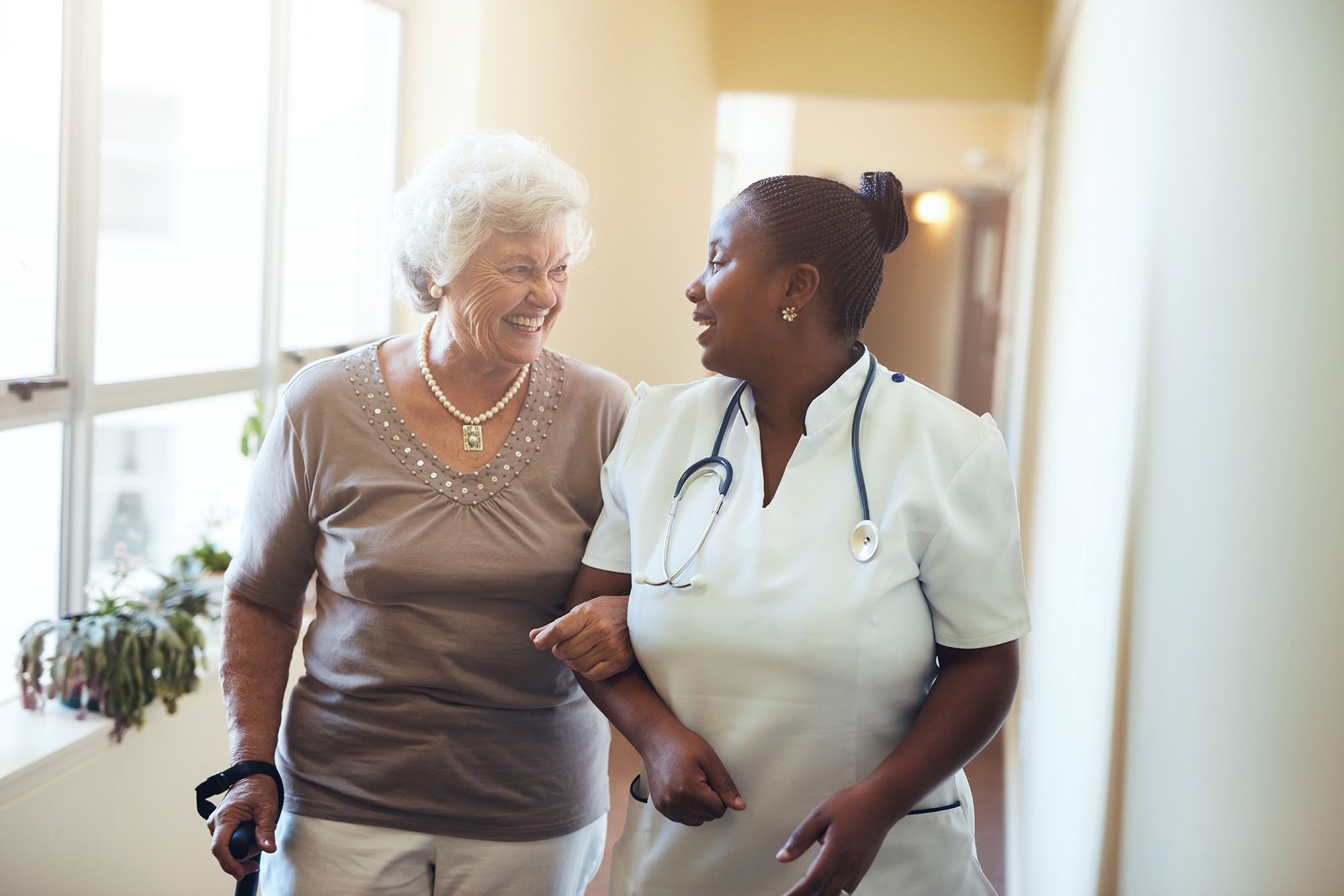 A smiling elderly woman walks arm-in-arm with a nurse in a hallway, highlighting supportive healthcare interactions and companionship.